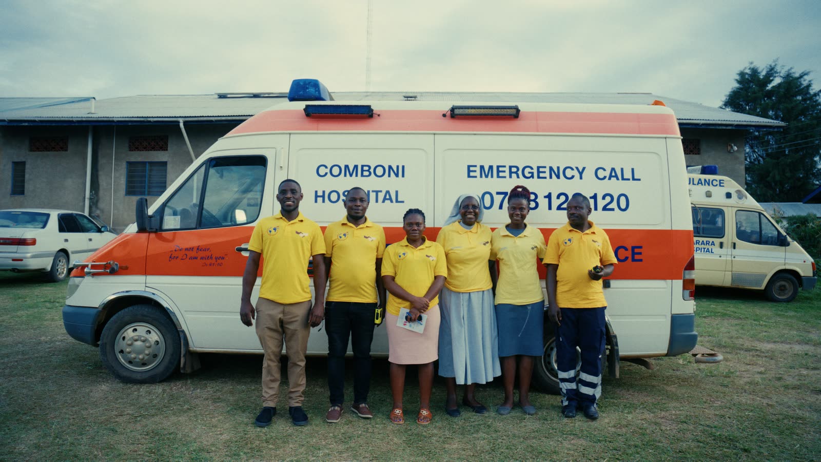 Das Team des Comboni Hospital in gelben Hemden vor seinem Krankenwagen mit Notrufnummer.